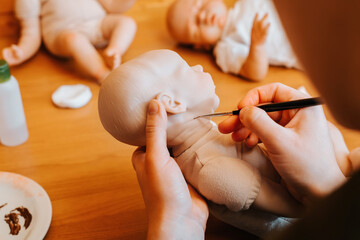 Close-up of female craftsman decorating with brush realistic doll of newborn caucasian child,...