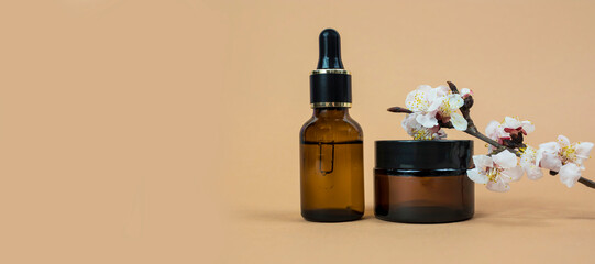 Glass serum bottle with pipette and cream jar with natural flowering branch on beige background.