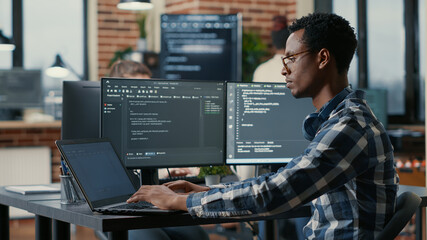 Portrait of african american developer using laptop to write code sitting at desk with multiple...