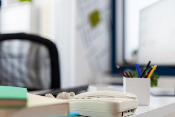 Closeup of phone on desk in empty business office. Startup workspace furniture in indoors work location. Modern commercial workplace design with telephone on white table with supplies.