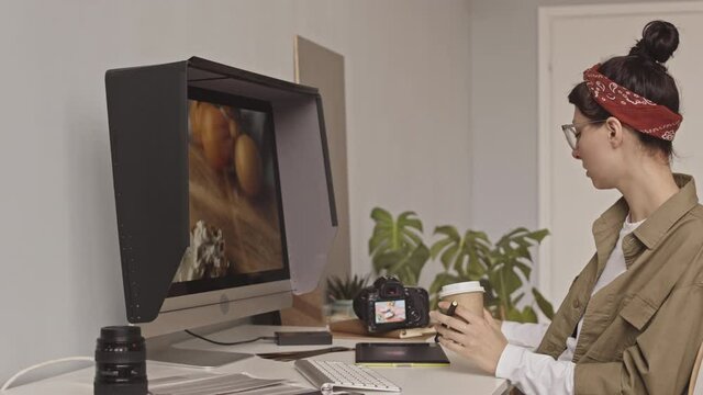 Waist up shot of young female photographer editing flat lay photos on computer using graphic tablet and stylus, sitting in her office