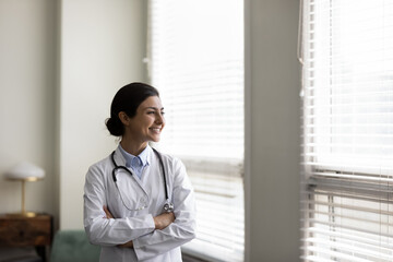 Fototapeta premium Confident happy young Indian woman GP doctor therapist physician cardiologist in white medical coat standing with folded arms in clinic office, looking in distance thinking of illness treatment.