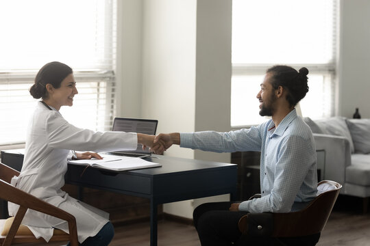 Smiling friendly young female Indian doctor shaking hands with African American male patient, getting acquainted or celebrating signing medical insurance agreement contract at meeting in clinic office