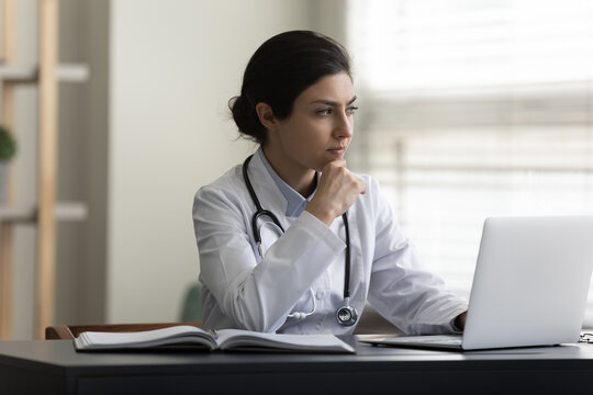 Stressed young Indian female doctor therapist in white medical uniform looking in distance, thinking of difficult problem solution, considering illness treatment working on computer in modern clinic.