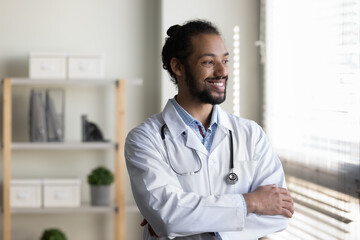 Happy dreamy young African American male doctor physician therapist in white coat standing with...