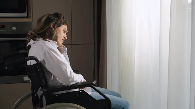 Pensive disabled woman with infantile cerebral paralysis sits in wheelchair near window behind curtain at home experiencing ableism side view.