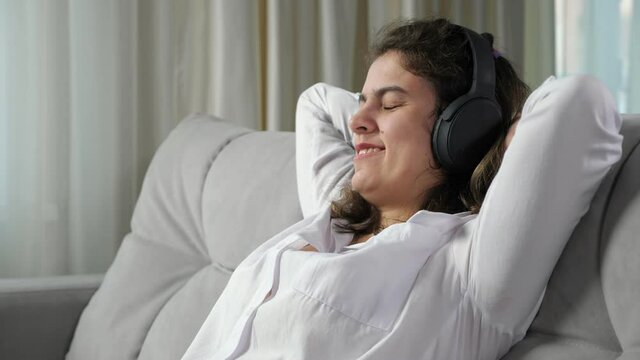 Young disabled long haired woman with cerebral palsy relaxes smiling and listening to music in headphones leaning back on sofa at home closeup.