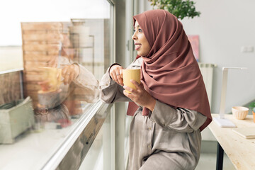 Contemplative woman with coffee cup looking through window at home