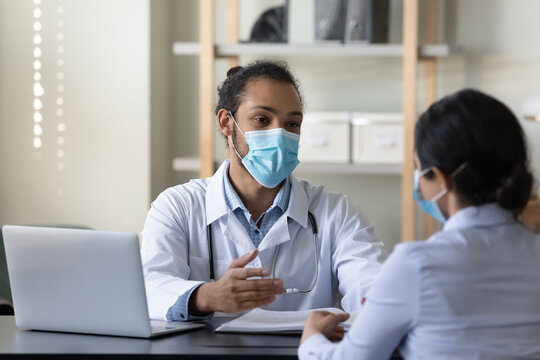 Happy friendly young African American male doctor therapist physician and female Indian patient in facial masks respirators communicating sitting at table in clinic office, medical healthcare concept.
