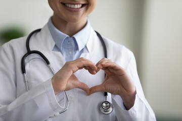 Close up happy young Indian female GP doctor therapist physician making heart gesture with fingers,...