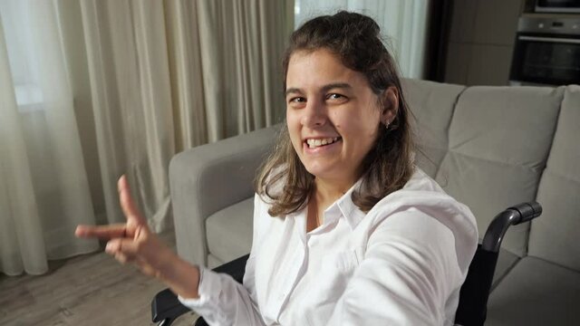 Disabled woman with cerebral palsy makes fingers Victory sign and smiles waving hand sitting in wheelchair expressing equality of people with disabilities.