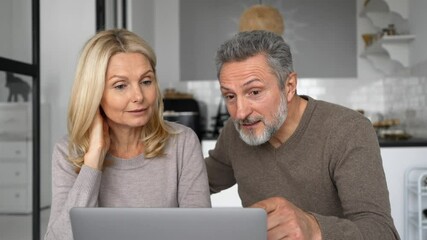 Cheerful middle-aged couple with a laptop sitting at the desk in modern kitchen at home, choosing new furniture, making food order, scrolling news feed. Mature spouses spend leisure time together - Powered by Adobe