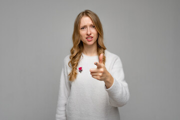 Fototapeta premium Concerned strict young blonde woman pointing index finger on camera choosing you. I blame you in all my problems. Girl wears white sweater, standing against gray background