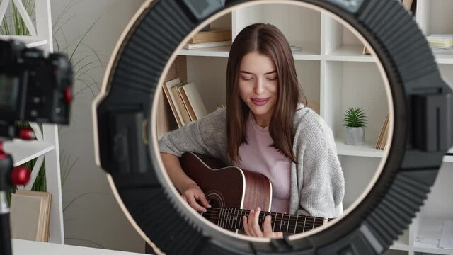 Female singer. Video record. Tutorial lesson. Inspired woman playing guitar on photo camera circle led lamp frame in light room interior.