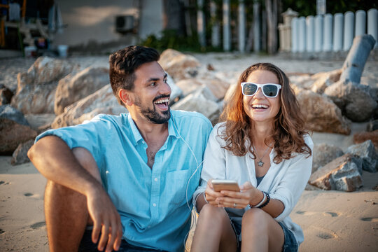 Happy beautiful young couple sitting on the beach on the sand with smartphone laughing and listening to music