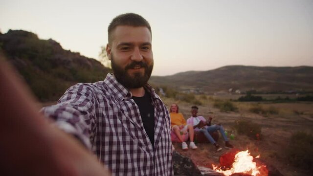 Cheerful man shoots himself on selfie camera and smiles. Bearded happy hipster holds camera and takes cool selfie. In background, two friends are sitting on bag chairs in camping outdoor