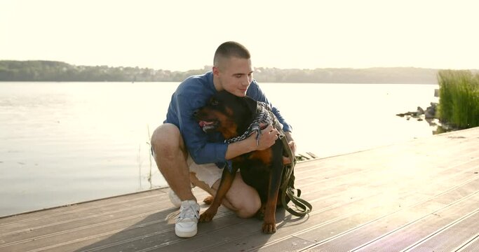 Young man stroking black rottweiler on lake pier