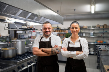 Confident chef and cook standing with arms crossed and looking at camera in commercial kitchen.