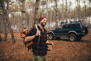 Millennial man hipster with beard wearing backpack, holding axe and firewood in forest with car on background.