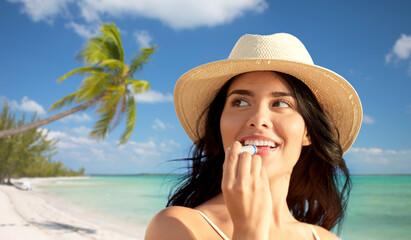 travel, tourism and summer vacation concept - portrait of happy smiling young woman in bikini swimsuit and straw hat applying lip balm over tropical beach background in french polynesia - obrazy, fototapety, plakaty