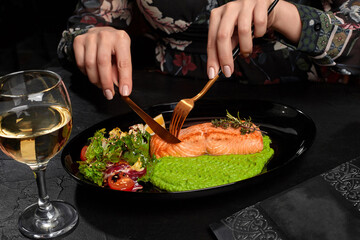 Female hands cutting grilled salmon steak served with pea puree, fresh salad, lemon and glass of wine