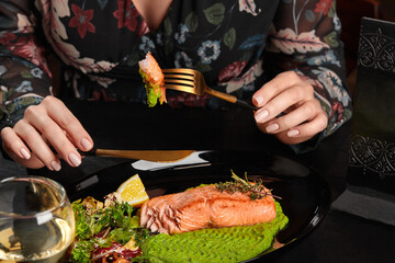 Female hands holding knife and fork with baked trout steak piece served with mashed peas, salad, lemon