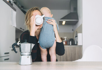 Modern tired mother and little child after sleepless night. Exhausted woman with baby is sitting with coffee in kitchen. Life of working mother with baby. Postpartum depression on maternity leave.