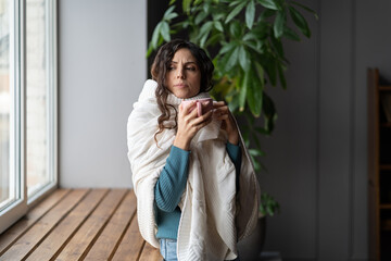 Young upset woman with sore throat feeling unwell drinking warm tea, standing by window at home, sick female having COVID infection symptoms fever, chills and cough. Hydration for flu recovery