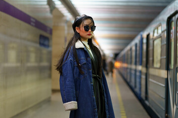 Obraz premium Stylish korean girl in eyeglasses and street fashion outfit at underground platform with approaching train. Trendy japanese tourist woman travel by public transport. Lifestyle and big city concept