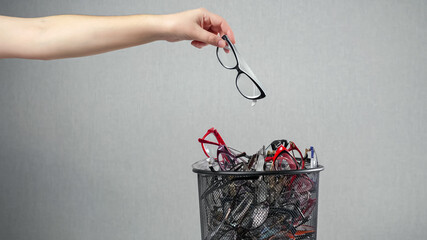 Woman throws out unnecessary glasses for eyesight on pile in rubbish bin against grey background as...