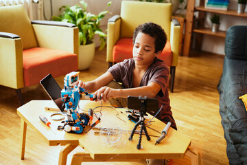A smart African-American boy holding soldering iron and touching tablet while making a robot at...