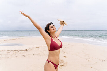 Cropped image of latin american woman on tropical island seashore with raised arms. Panoramic view of woman in bikini with hat on her hand on paradisiacal beach. People traveling and holidays concept