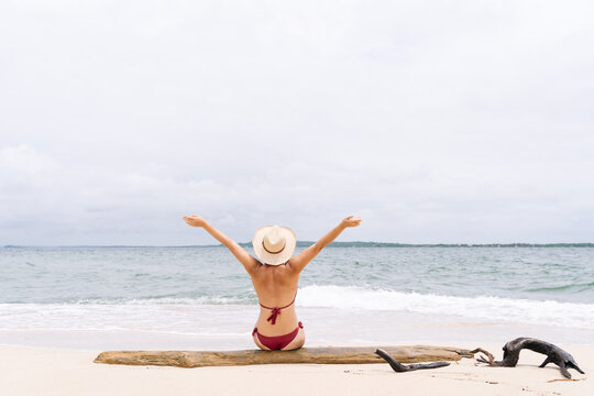 Full length image of unrecognizable woman on tropical island seashore with raised arms. Panoramic rear view of woman in bikini with hat on paradisiacal beach. People traveling and holidays concept
