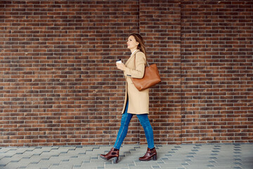 Fototapeta na wymiar A woman in a coat walking and holding takeaway coffee. A young fashionable woman walking on the street, passing by a brick wall and holding a hot beverage in a disposable cup.