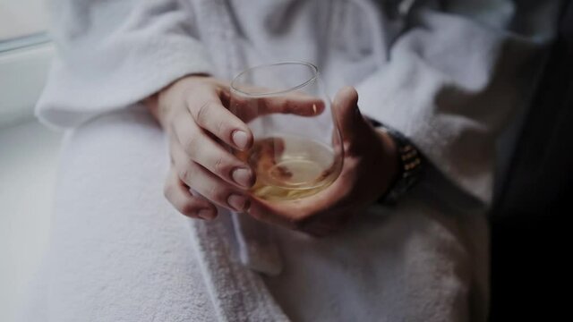 Man in a bathrobe holding a glass of whiskey sitting on the windowsill. The man drinks alcohol.