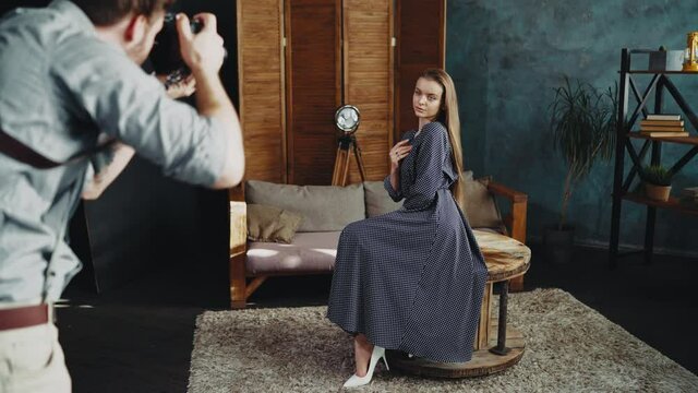 A young pretty sexy model with long hair in a long dress poses for a photographer sitting on a table in an interior loft studio