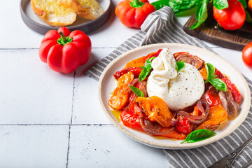 Burrata cheese with baked tomatoes, pepper, red onion and fresh basil on white tile background. Traditional italian food.