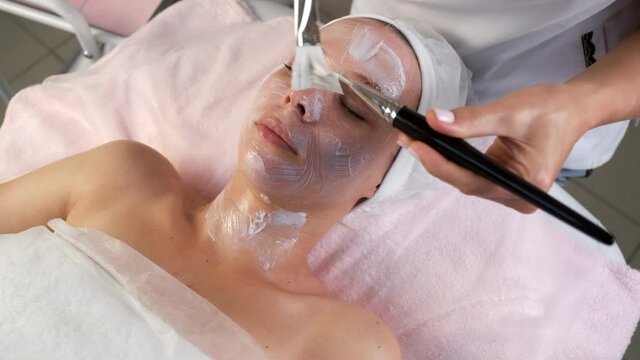 Professional beautician is applying mask to woman's face using two brushes in cosmetology clinic. Cosmetologist doctor is making beauty procedure against facial wrinkles. Anti-aging effect.