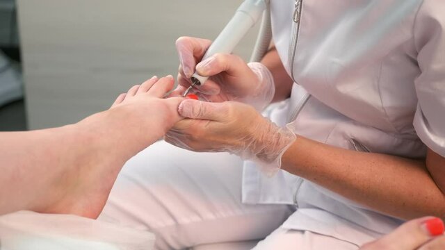 Pedicurist woman removes shellac polish from toes using manicure machine, closeup view. Pedicure Master is removing red gel polish from nails on toes using electric nail drill in cosmetology.