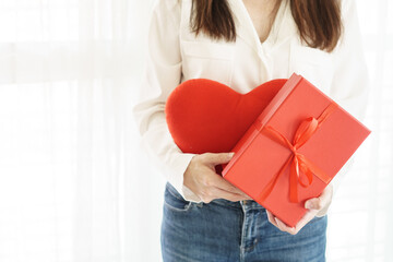 Woman in white shirt and jean holding red heart pillow and red box present with red ribbon, white curtain background, space on left
