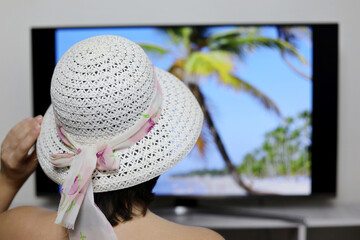 Woman in sun hat looking at TV screen with tropical beach and coconut palm trees. Girl dreaming about sea holidays, planning summer travel