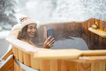 Happy woman relaxing in hot bath outdoors, talking on phone while sitting in thermal spa at snowy...