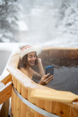 Happy woman relaxing in hot bath outdoors, talking on phone while sitting in thermal spa at snowy...