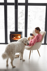 Woman sitting with her white dog by the fireplace at modern house on nature during winter time....