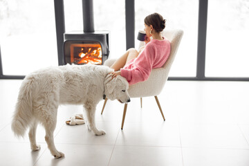 Woman sitting with her white dog by the fireplace at modern house on nature during winter time. Concept of winter mood and comfort at home. Idea of friendship with dog