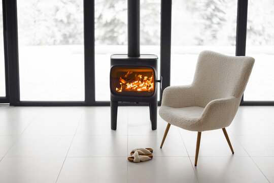 Cozy living space by the burning fireplace with chair, cup and slippers on background of snowy landscape. Idea of home comfort and winter mood