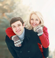 Portrait of happy smiling couple in winter park, man giving piggyback ride to woman on snowy forest...