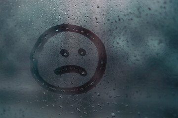 Sad smiley drawn by hand on wet fogged glass from the rain, copy the space. Concept of loneliness, sadness, bad weather