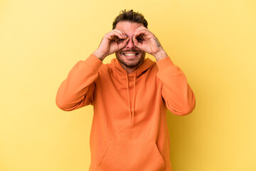 Young caucasian man isolated on yellow background showing okay sign over eyes