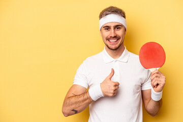 Young caucasian man holding ping pong rackets isolated on yellow background smiling and raising...